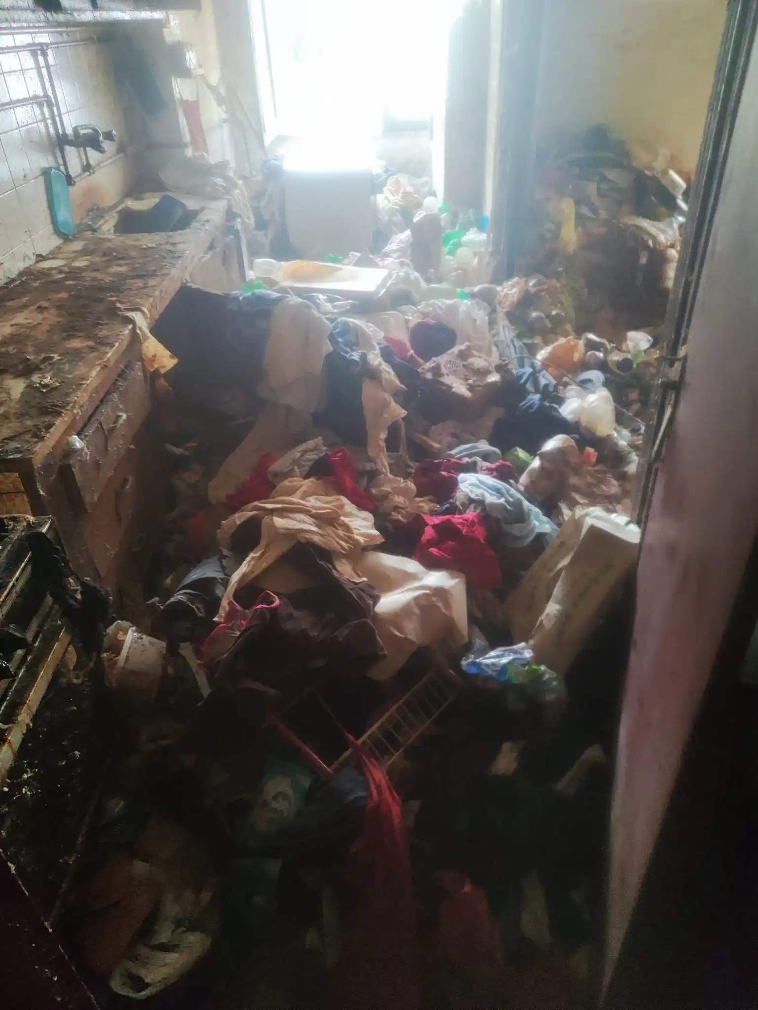 Cozinha em estado de acumulação extrema: bancadas cobertas por camadas de gordura escura e resíduos secos, fogão com queimadores obstruídos por crostas negras carbonizadas, lava-louças transbordando de louça suja empilhada, chão pegajoso com manchas escuras espalhadas e lixo acumulado nos cantos.